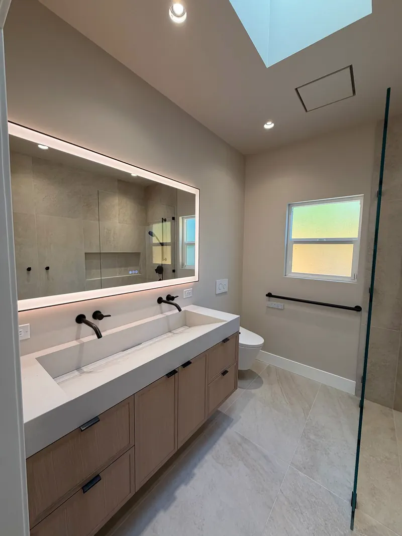 Bathroom with a floating wood vanity, wide trough sink, wall-mounted black faucets and a backlit mirror